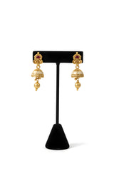 Close up of gold plated trefoil stud jhumka earrings with ruby accent white enamel dome and gold ball drop